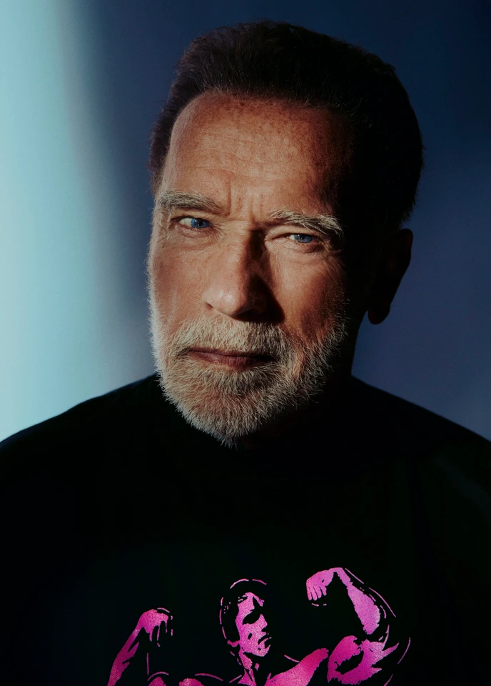 Actor Arnold Schwarzenegger, steel gaze and silver beard, in a black bodybuilder graphic tee against a cool blue gradient.