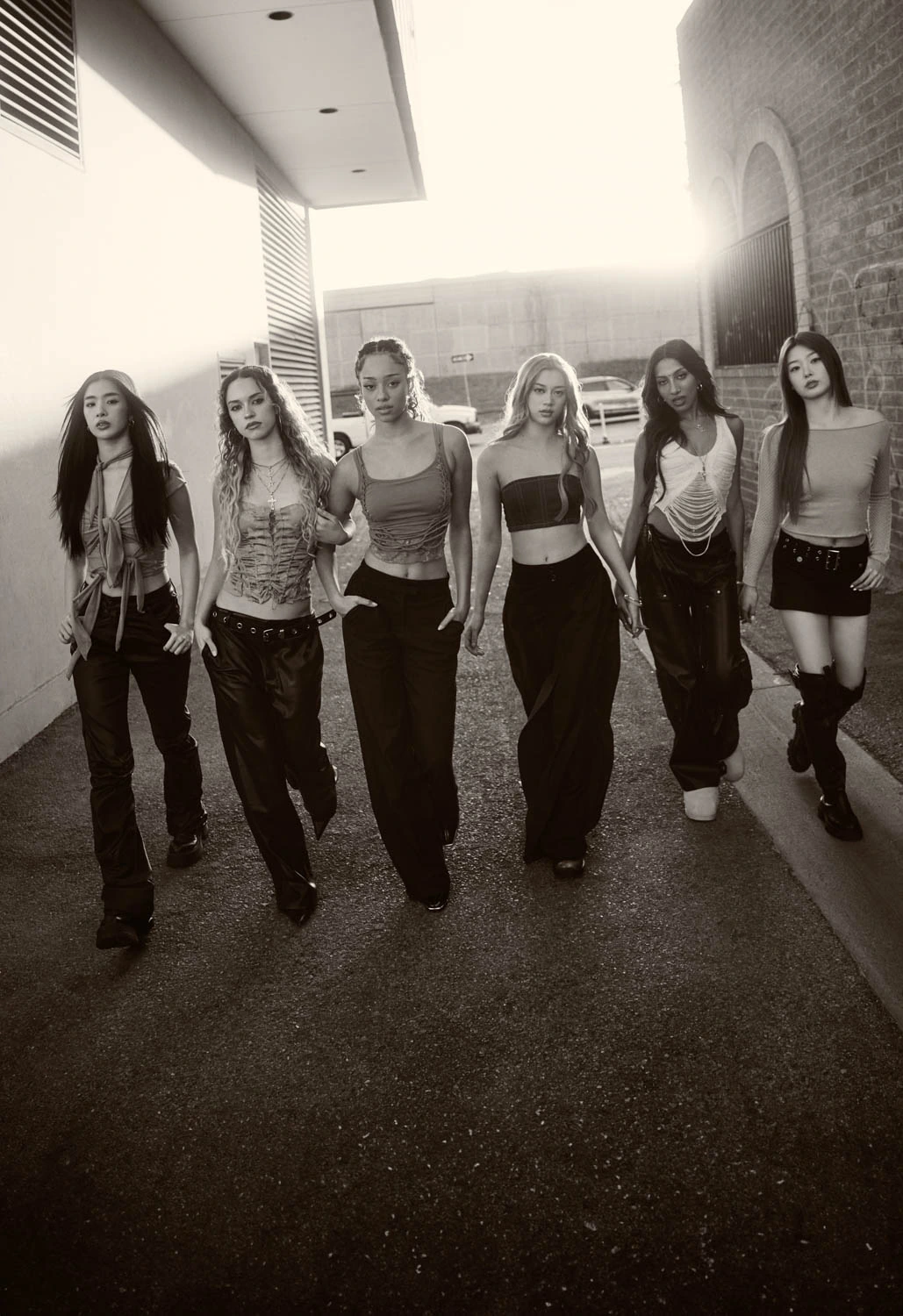 Girl group Katseye — all six members — stride an alley in black-and-white, crop tops and wide-leg pants, backlit haze behind.