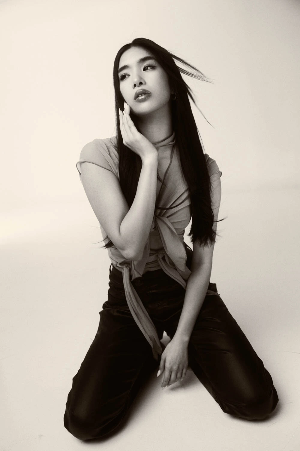 Katseye's Sophia kneeling on a white studio floor, tied mesh top and leather trousers, hand raised to cheek, black-and-white.
