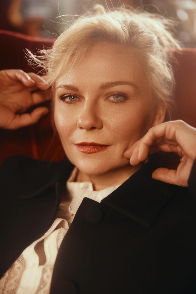 Actor Kirsten Dunst, blonde updo and deep red lip, black blazer over ivory lace, hands raised, warm amber light.