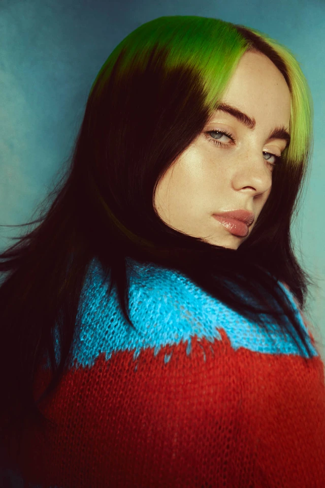 Pop star Billie Eilish, green-black hair, bold red-blue knit sweater, glancing over shoulder on blue, by Austin Hargrave