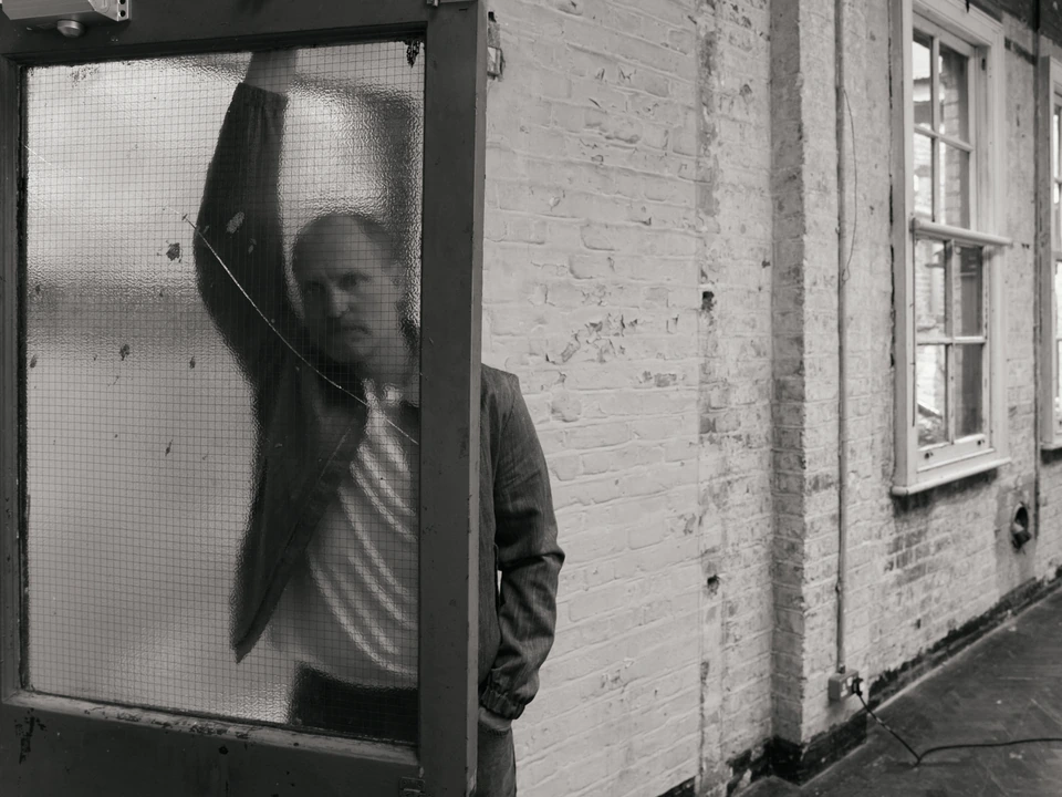 Actor Woody Harrelson half-obscured behind frosted wire glass, arm raised, brooding in a dark jacket against a worn brick.