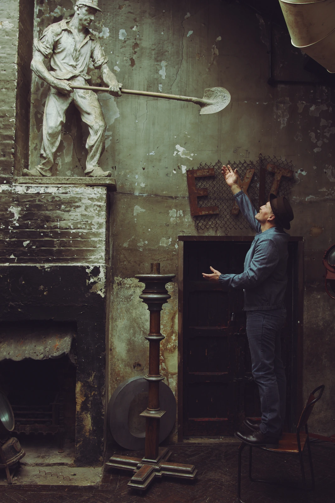 Actor Woody Harrelson arms raised toward a laborer statue in a decayed industrial loft, moody green light, denim jacket.
