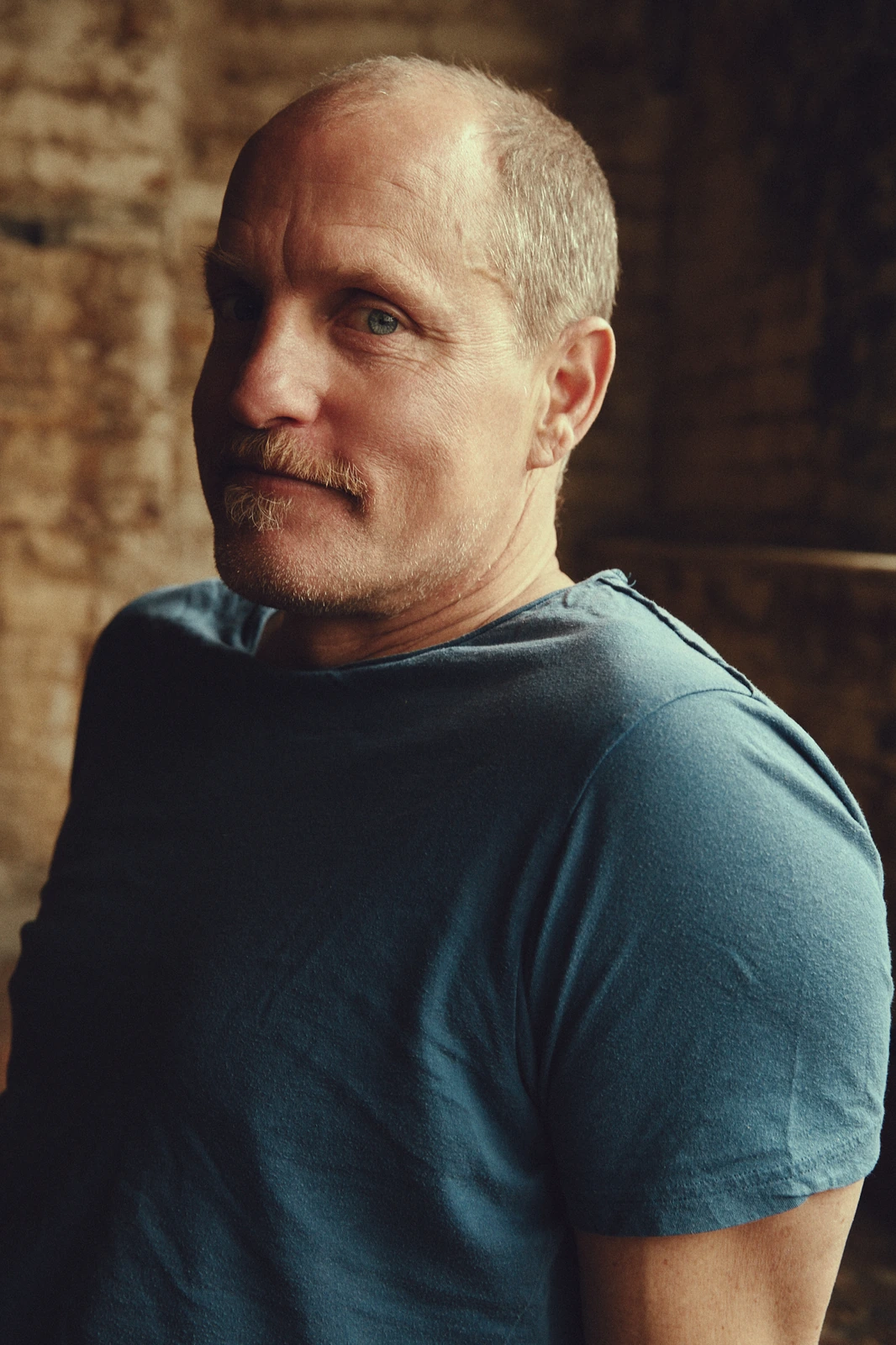 Actor Woody Harrelson in a slate blue tee, gaze drifting off-frame against a warm raw-brick wall, for The Hollywood Reporter.