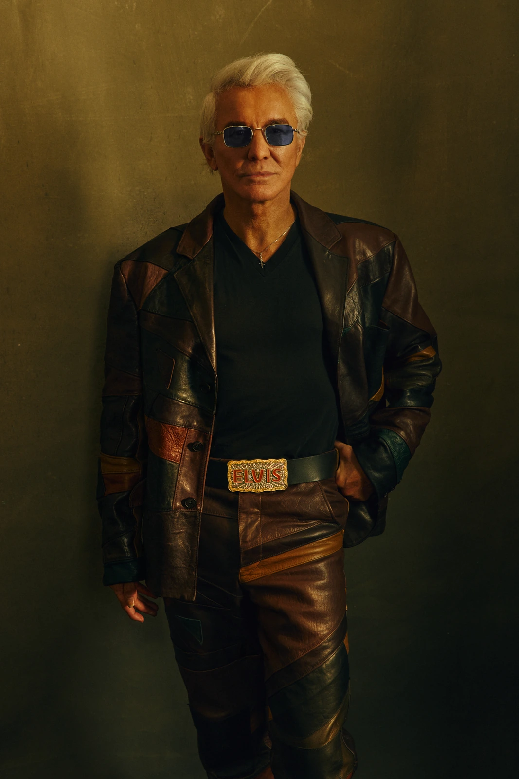 Director Baz Luhrmann in a patchwork leather jacket and ELVIS belt buckle, silver hair, blue shades, warm golden studio.