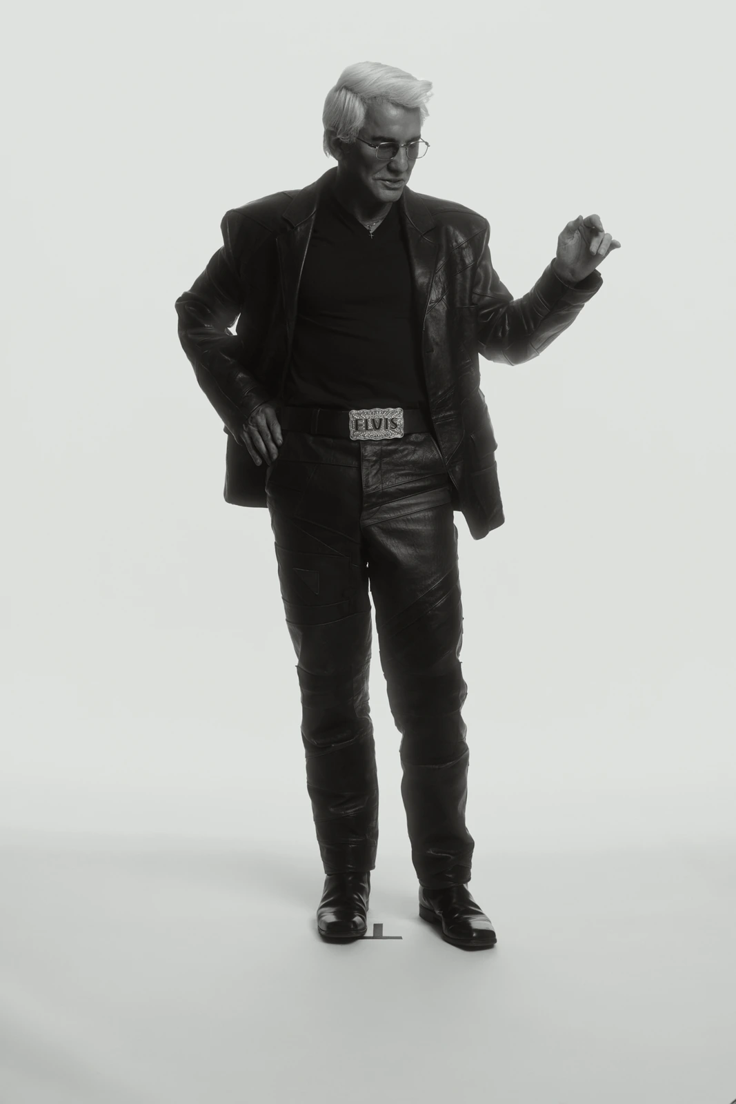 Director Baz Luhrmann in full black leather and an Elvis belt buckle, glancing down mid-gesture against a clean grey ground.