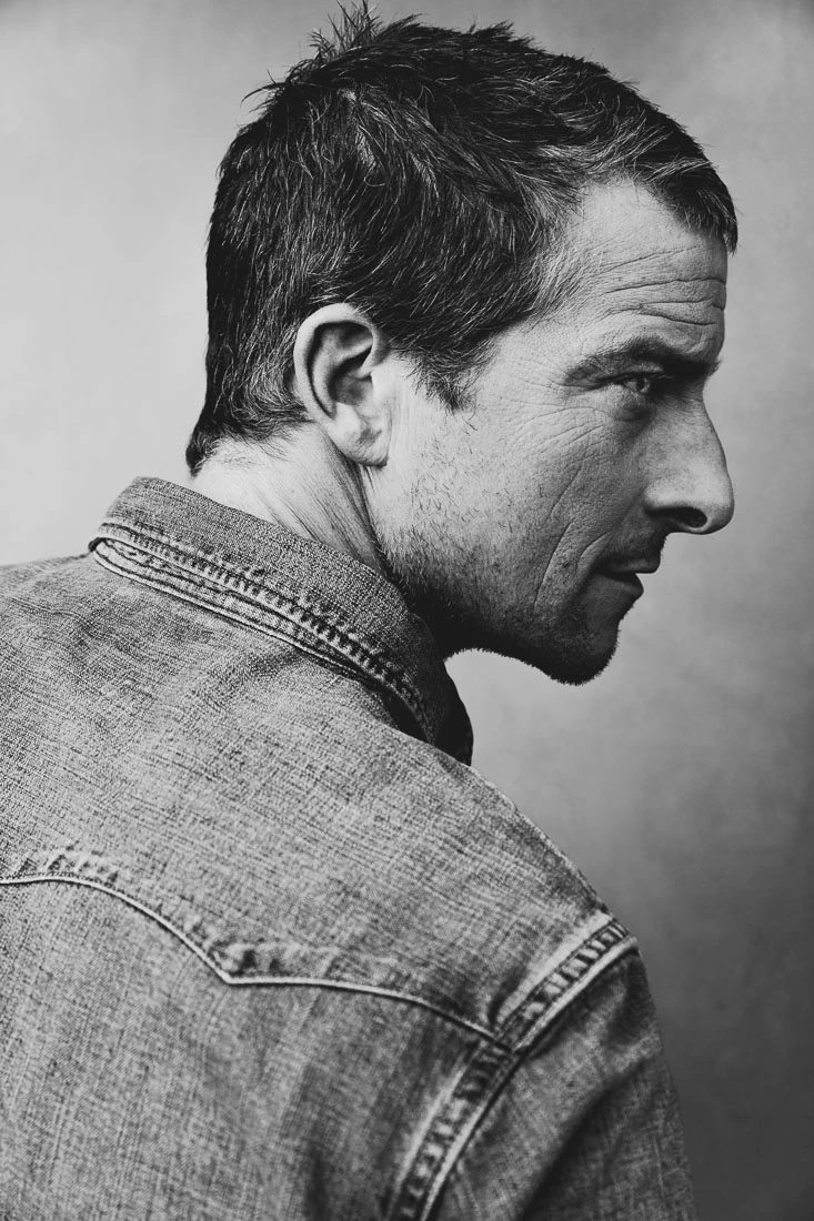 Adventurer and TV presenter Bear Grylls shot from behind in a denim jacket, sharp profile turned, black and white studio.