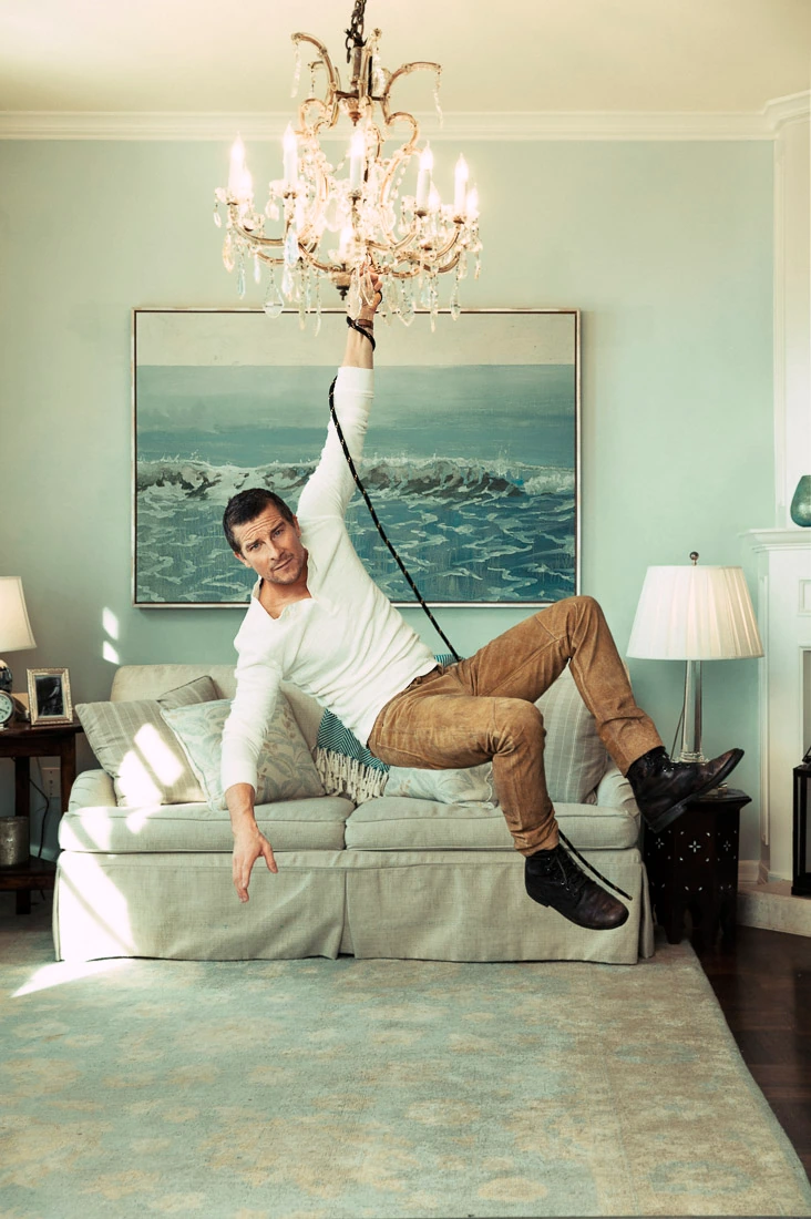Adventurer and TV presenter Bear Grylls swings from a chandelier over a sofa in a hotel living room, white top and tan.