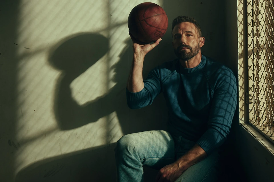 Actor and director Ben Affleck holds a worn basketball against a shadowed wall, striped light raking across his teal sweater.