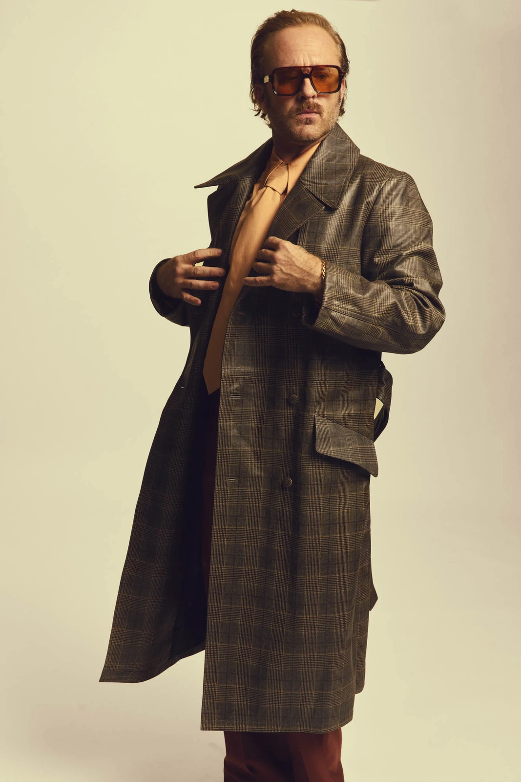 Actor Ben Foster in an oversized plaid trench coat, amber shades, open chest, cool gaze against a warm cream studio backdrop.