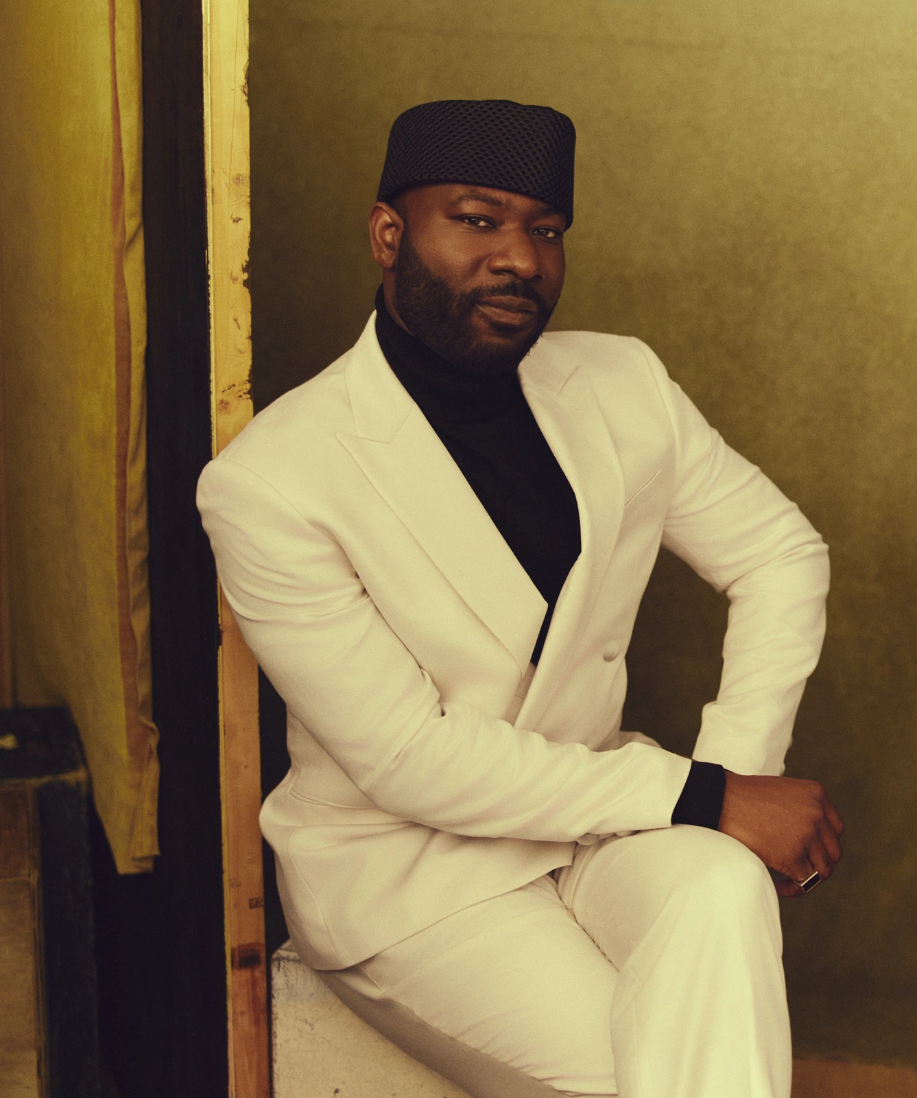 Director Blitz Bazawule seated in a white suit and black turtleneck, navy cap, warm olive backdrop with golden light.