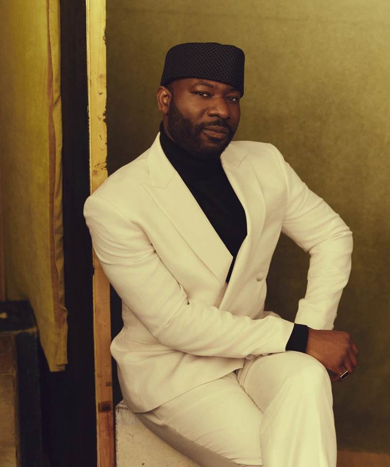 Director Blitz Bazawule seated in a white suit and black turtleneck, navy cap, warm olive backdrop with golden light.
