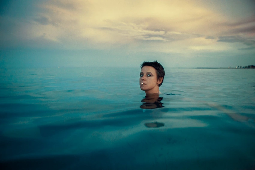 Musician Cat Power submerged to the chin in still open water, gaze drifting sideways beneath a wide dusk sky, cyan and gold.