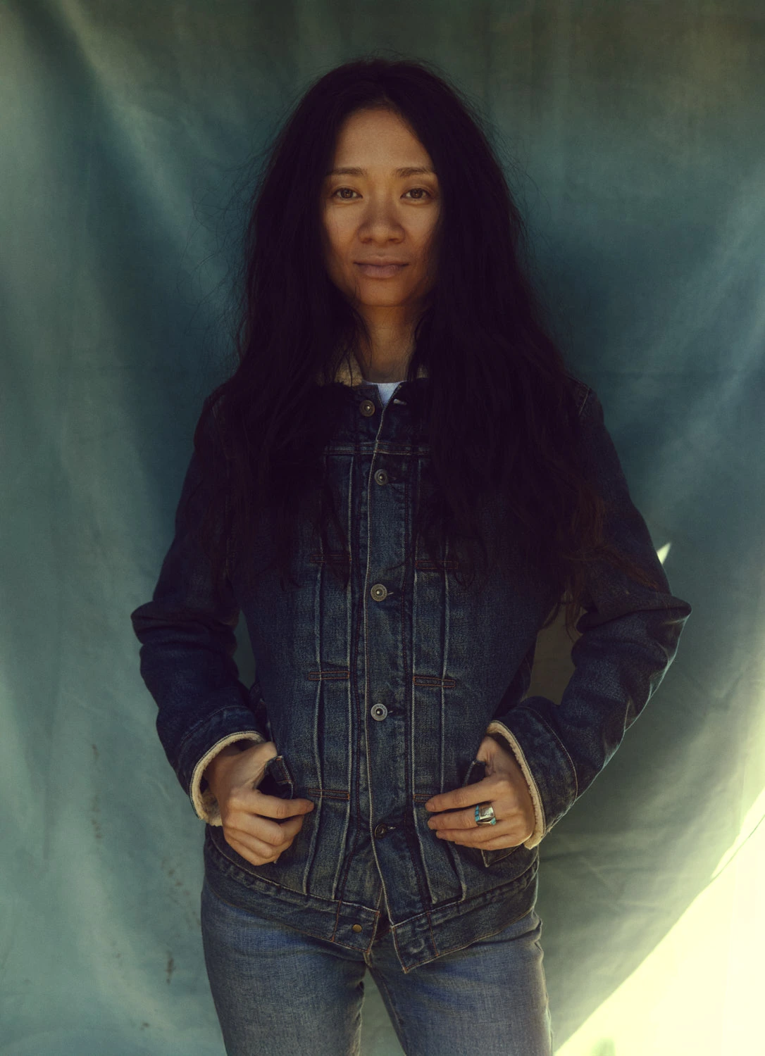 Director Chloé Zhao, Nomadland Oscar winner, in denim jacket hands in pockets, steady gaze against a teal drape, by Austin.