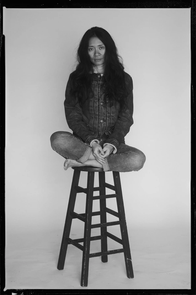 Filmmaker Chloé Zhao, Nomadland director, cross-legged on a stool in denim, steady gaze, B&W studio portrait by Austin.