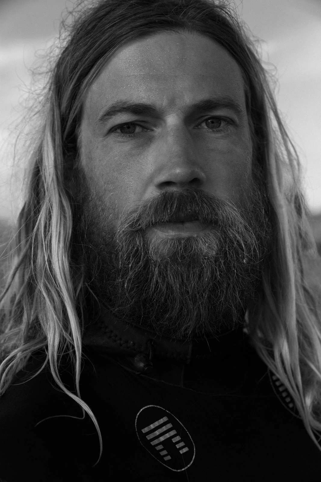 Chris Del Moro in a black-and-white close portrait, long blond hair, full beard, wetsuit logo visible, gaze steady and raw.