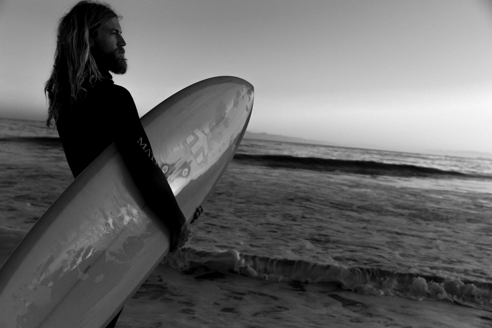 Chris Del Moro in a wetsuit, surfboard tucked under his arm, gazing toward the ocean at dusk in stark black and white.