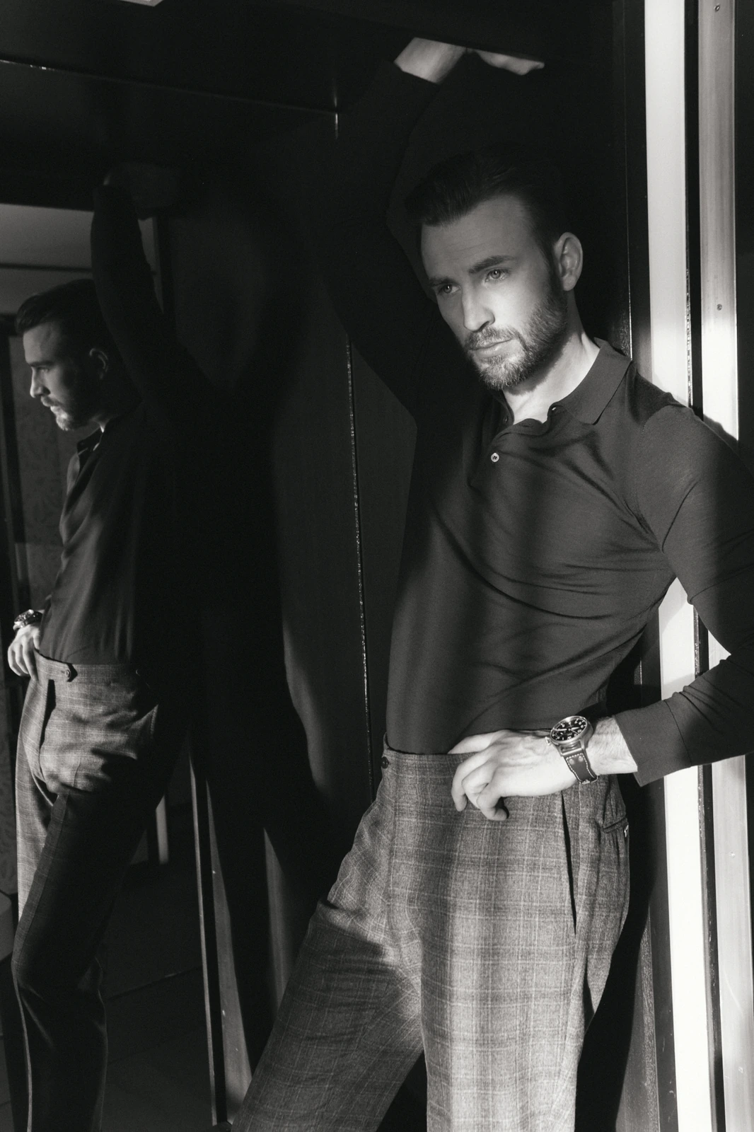 Actor Chris Evans leaning in a doorway, reflected in dark glass, tailored plaid trousers and polo, steel watch, black.
