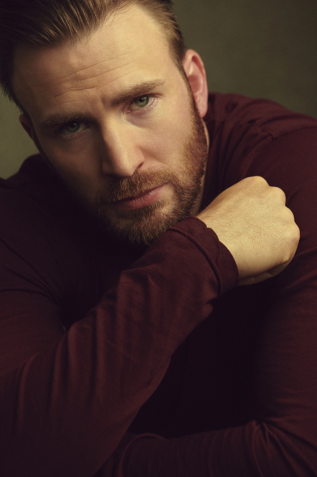 Actor Chris Evans in deep burgundy, arms crossed and gaze fixed, lit in warm shadow against a moody olive backdrop.
