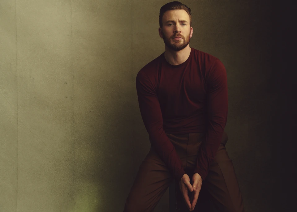 Actor Chris Evans seated and still in a deep red long-sleeve against a mossy green wall, hands clasped, warm shadowed light.