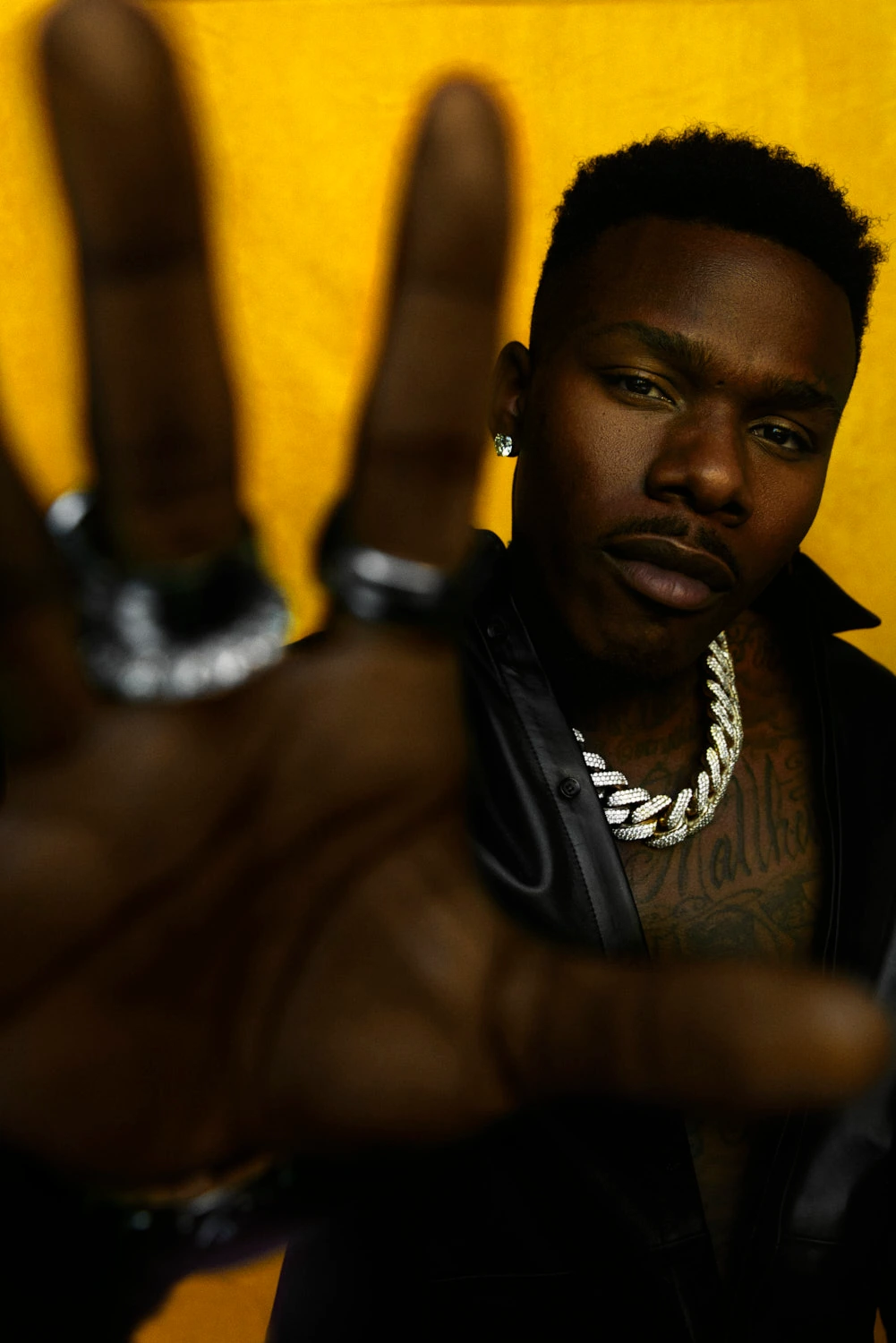 Rapper DaBaby throws a peace sign toward the lens, iced Cuban chain and chest tattoos against a bold yellow backdrop.