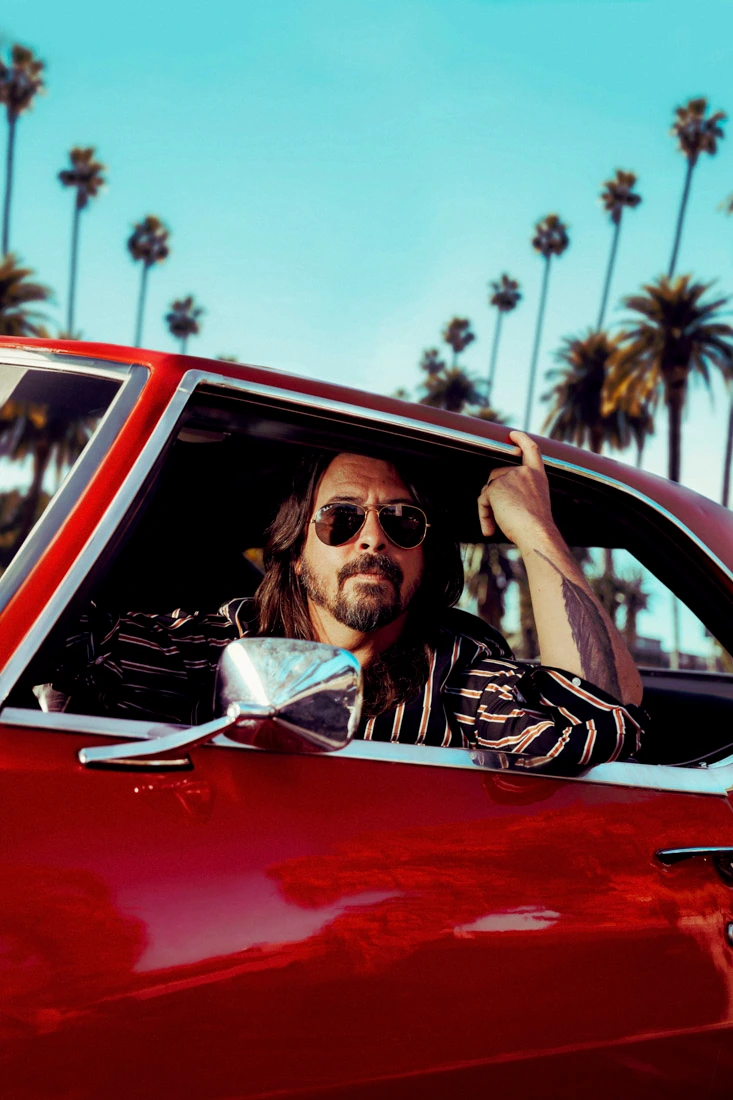 Musician Dave Grohl, Foo Fighters founder, leaning from a red classic car in aviators and a stripe shirt, palm trees.