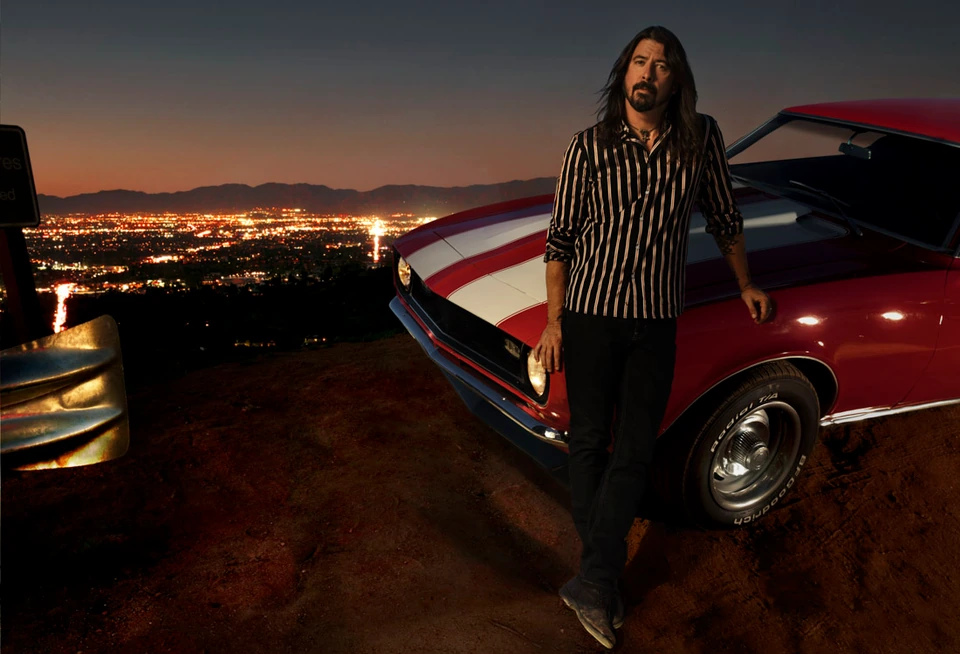 Musician Dave Grohl, Foo Fighters founder, leaning on a red classic muscle car at dusk, LA sprawling bright below.