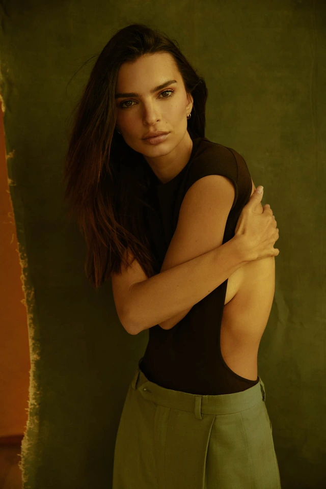 Model and actress Emily Ratajkowski, arms crossed in a black bodysuit and olive trousers, warm amber light against a deep.