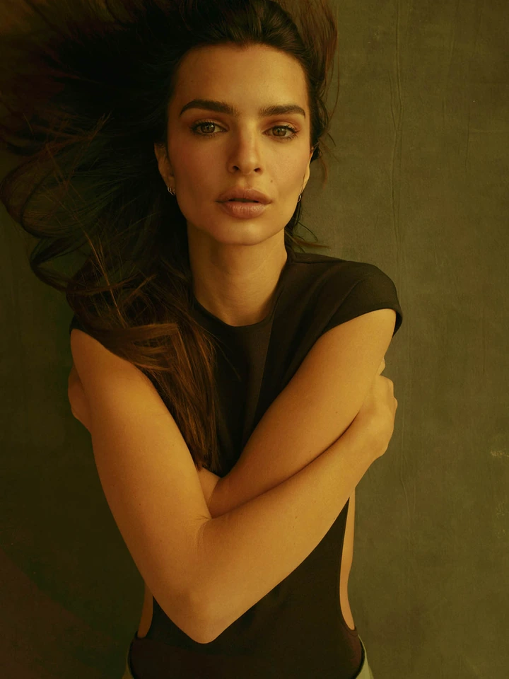 Model and actress Emily Ratajkowski, arms crossed, hair swept, in a dark cut-out top against warm olive, by Austin Hargrave.