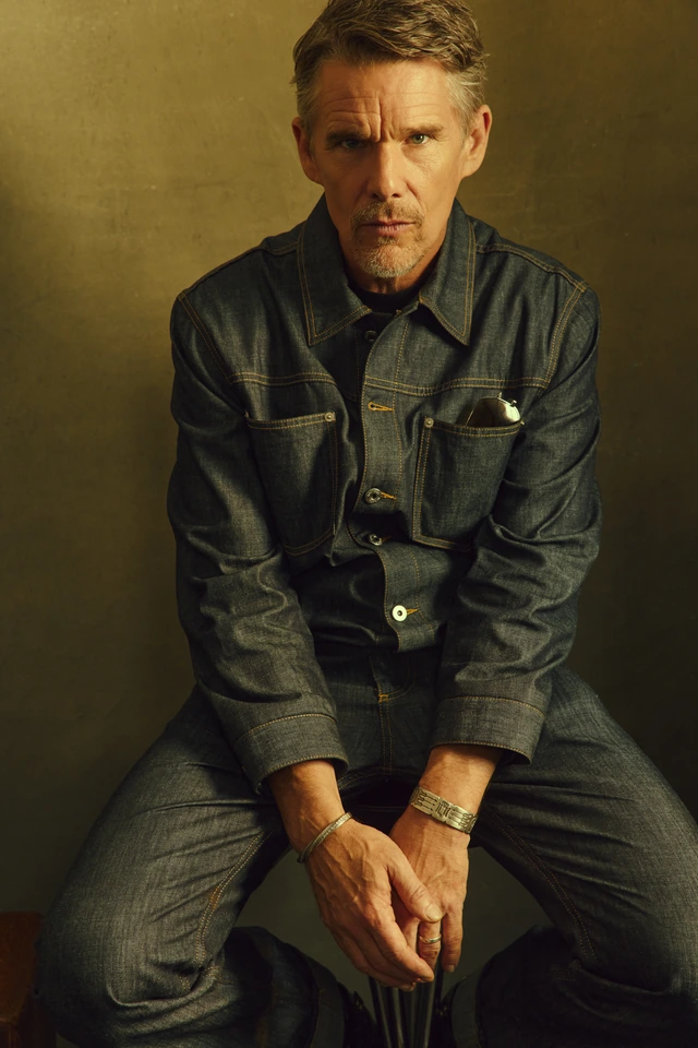 Actor Ethan Hawke seated in a dark denim jacket over jeans, hands clasped, steady gaze against a warm ochre backdrop.