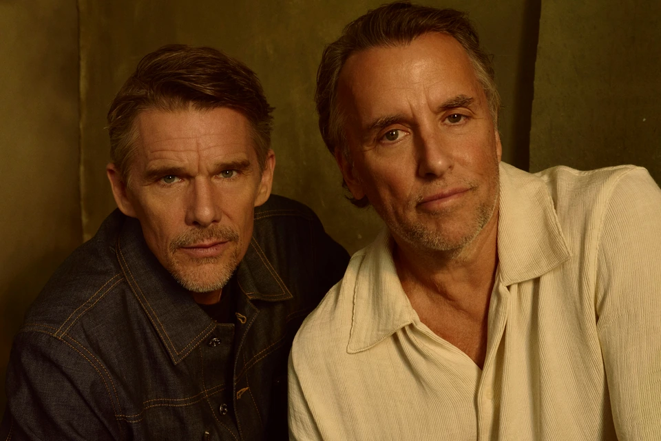 Actor Ethan Hawke and director Richard Linklater lean close, intense gaze, denim and cream against warm olive shadow.