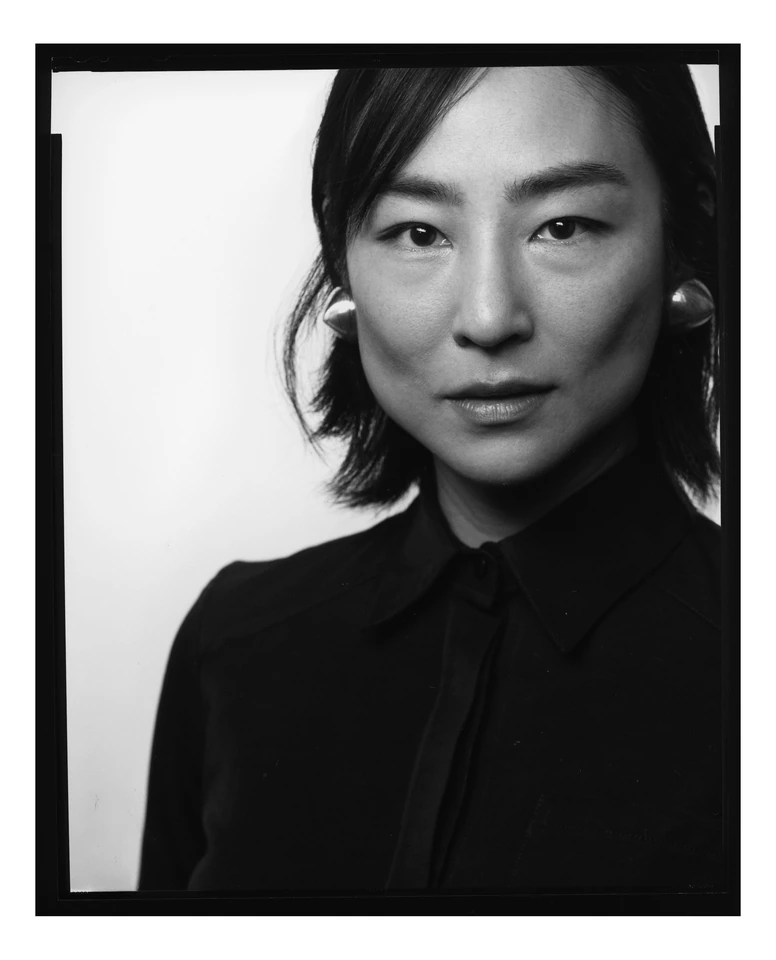 Actor Greta Lee in a black-and-white large-format portrait, dark button-up, pearl earring, steady gaze against soft white.