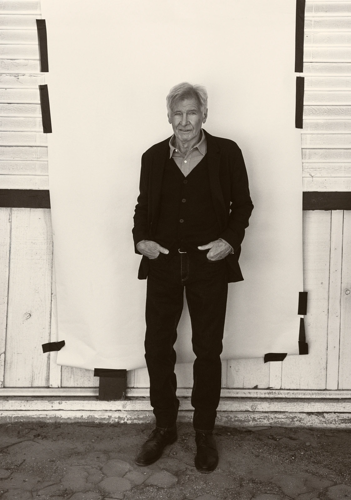 Actor Harrison Ford, hands in pockets, in a black blazer and open-collar shirt before a taped white backdrop, B&W outdoor.