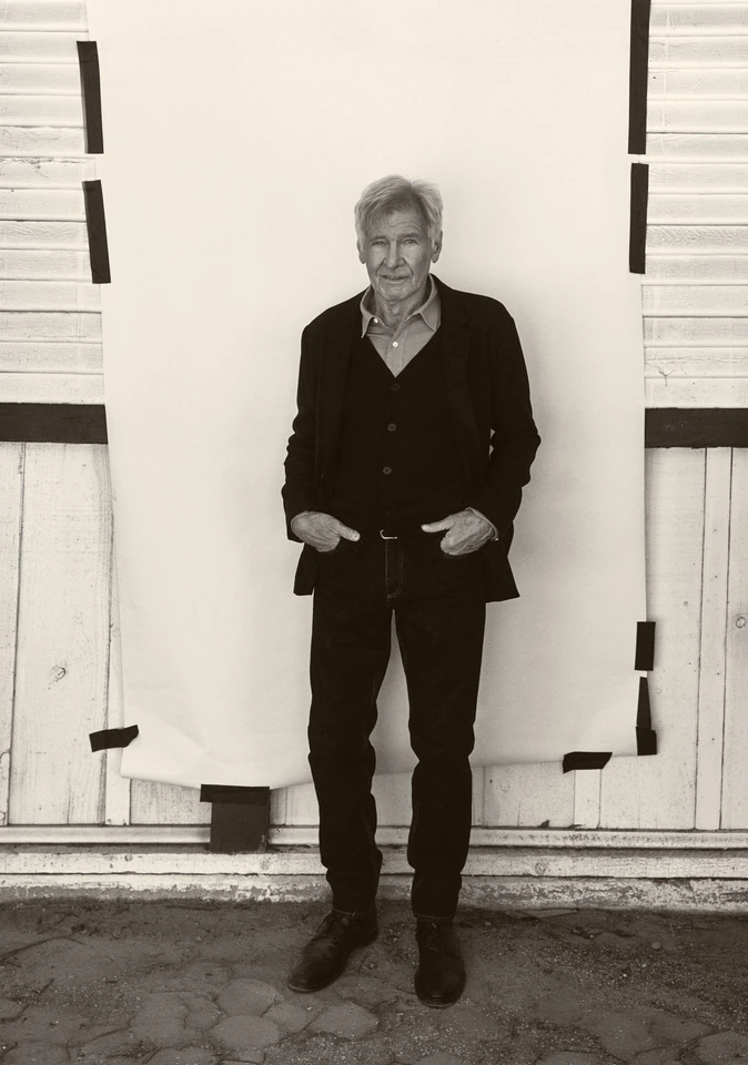 Actor Harrison Ford, hands in pockets, in a black blazer and open-collar shirt before a taped white backdrop, B&W outdoor.