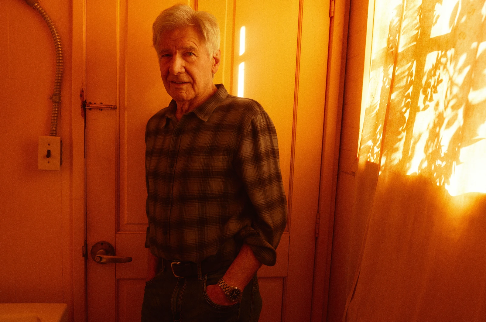 Actor Harrison Ford, hands in pockets, in a plaid flannel shirt by a wooden door, amber window light flooding the frame.