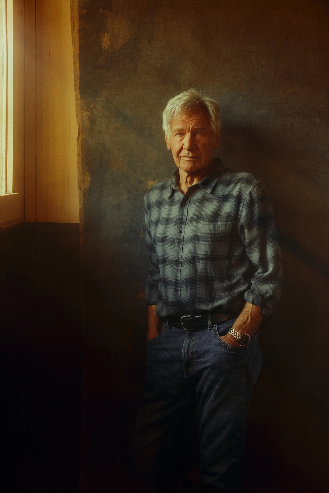 Actor Harrison Ford in a plaid flannel shirt and jeans, hands in pockets, leaning against a textured wall in warm raking.