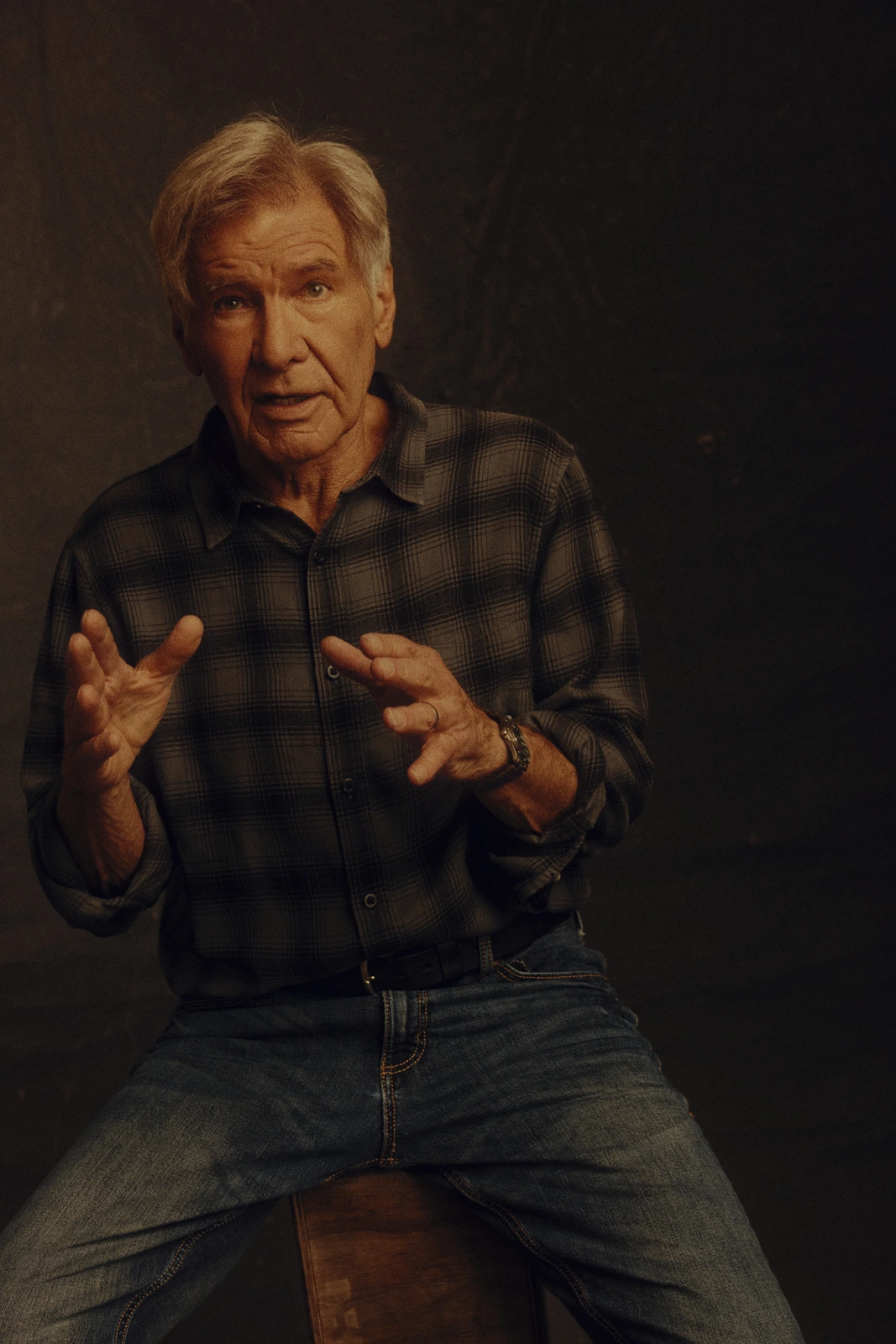 Actor Harrison Ford mid-gesture, hands open toward camera, in a plaid flannel shirt and jeans, warm amber studio light.