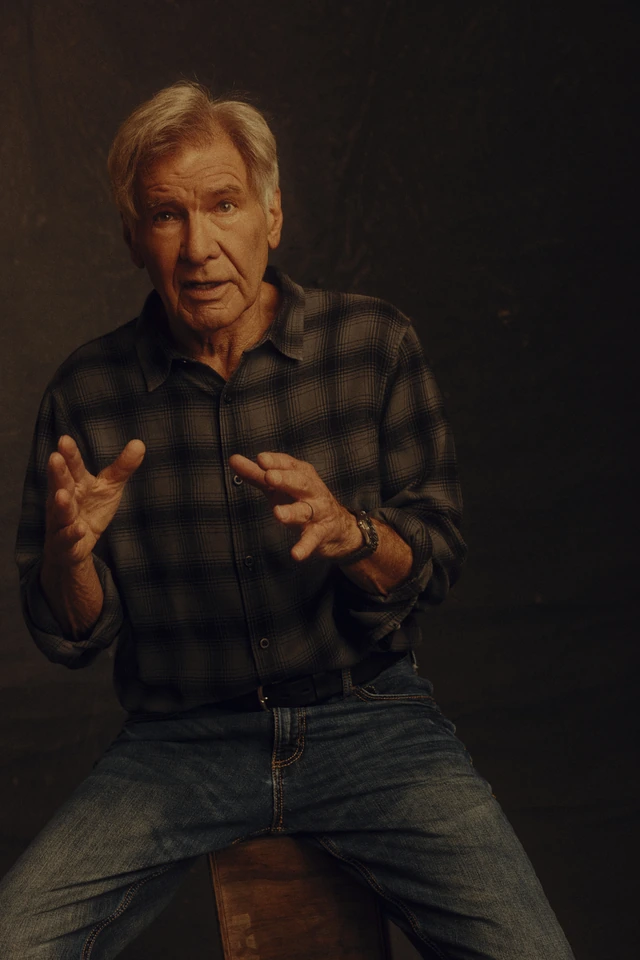 Actor Harrison Ford mid-gesture, hands open toward camera, in a plaid flannel shirt and jeans, warm amber studio light.