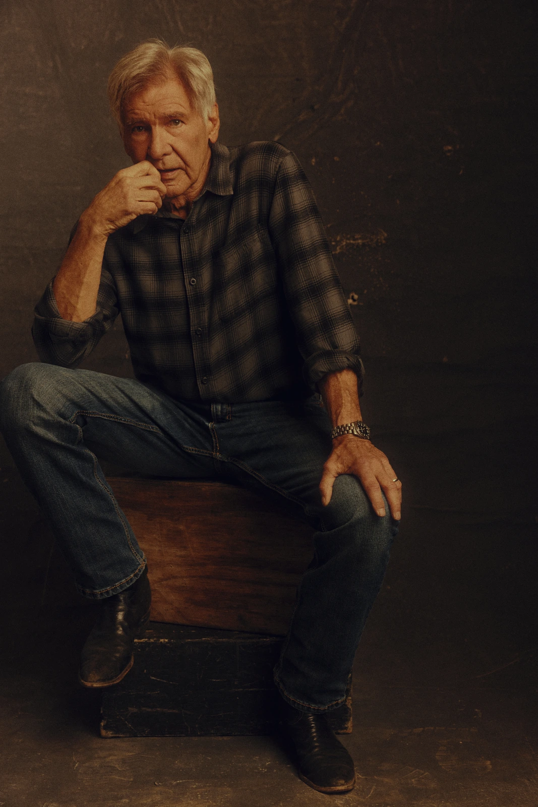 Actor Harrison Ford seated on a worn wooden crate, plaid flannel shirt and jeans, fist raised to his chin in quiet thought.