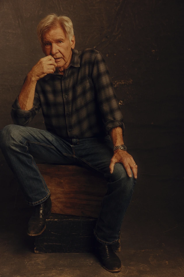 Actor Harrison Ford seated on a worn wooden crate, plaid flannel shirt and jeans, fist raised to his chin in quiet thought.