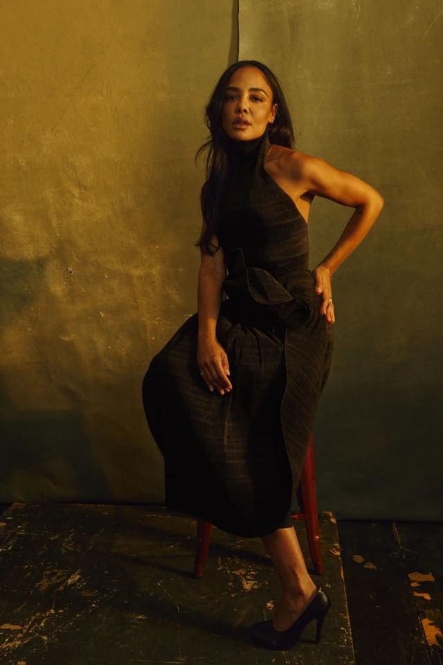 Actor Tessa Thompson in a sculptural black halter midi dress, hand on hip against a warm gold-washed studio wall, amber.