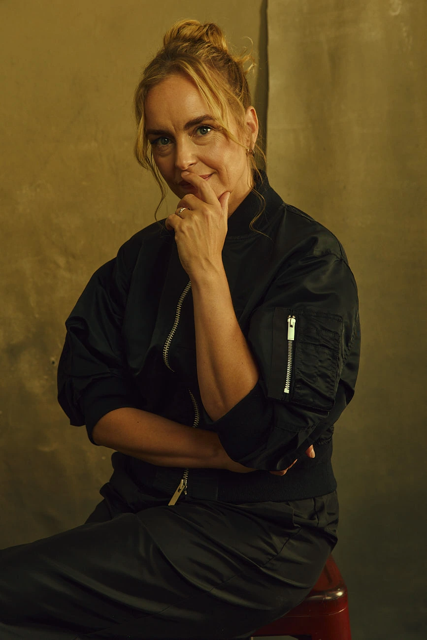 Actor Nina Hoss in a black bomber jacket, seated with a wry smile and hand at chin against a warm ochre backdrop, by Austin.