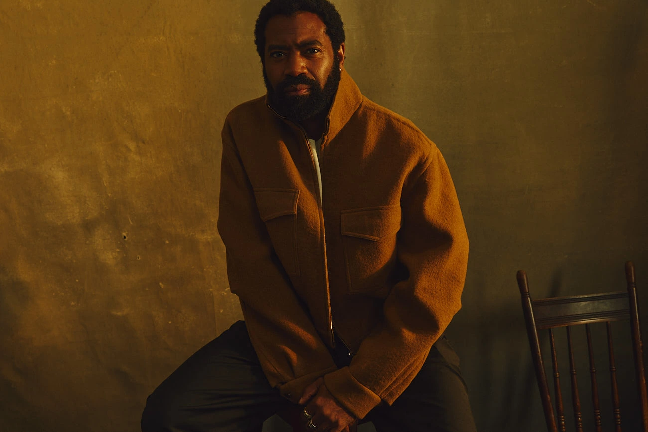 Actor Nicholas Pinnock seated in a caramel wool jacket, hands clasped, steady gaze against a warm ochre-washed wall.