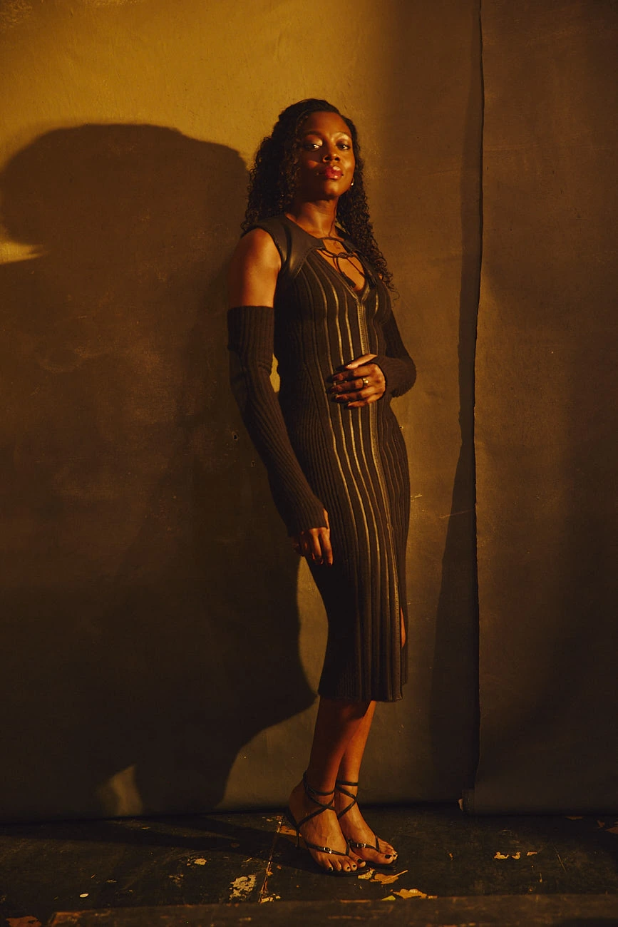 Director Nia DaCosta in a ribbed knit dress and arm warmers, leaning against a warm amber wall, calm gaze, golden light.