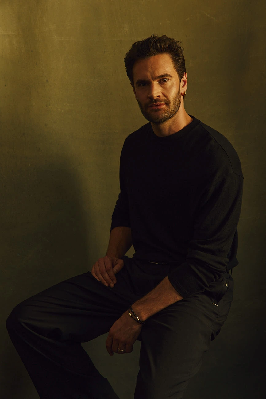 Actor Tom Bateman perched on a stool in a black long-sleeve, dark trousers, direct gaze against olive-washed warmth.