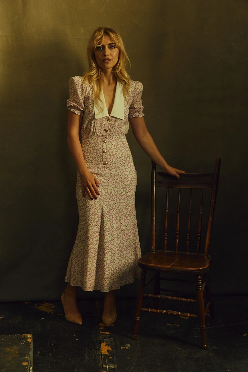 Actor Imogen Poots in a floral midi dress with white sailor collar, hand resting on a wooden chair, warm amber studio light.