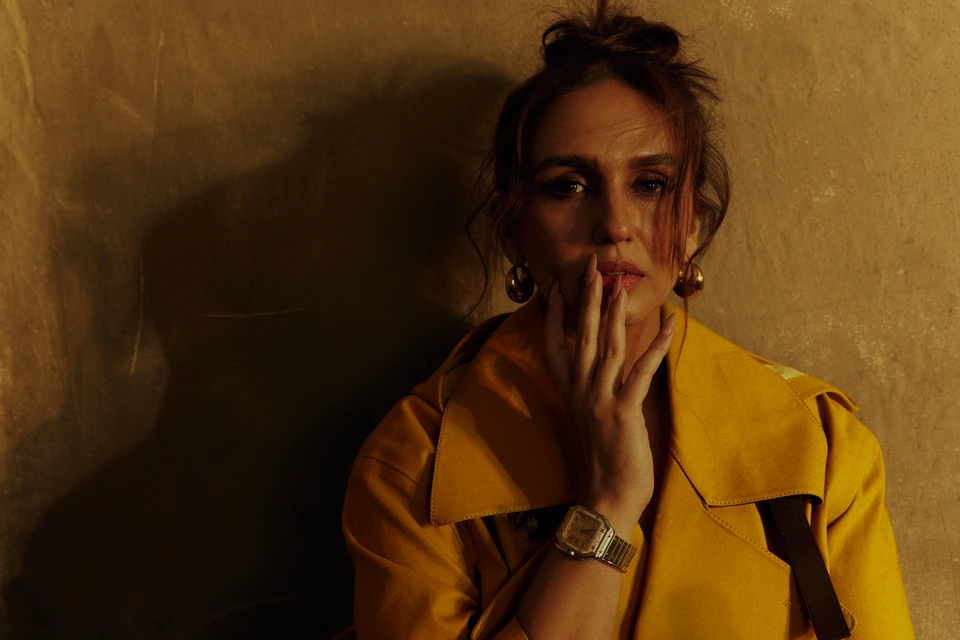 Actress Huma Qureshi of Maharani in a mustard trench, gold hoops, hand raised to lips against a smoldering amber wall.