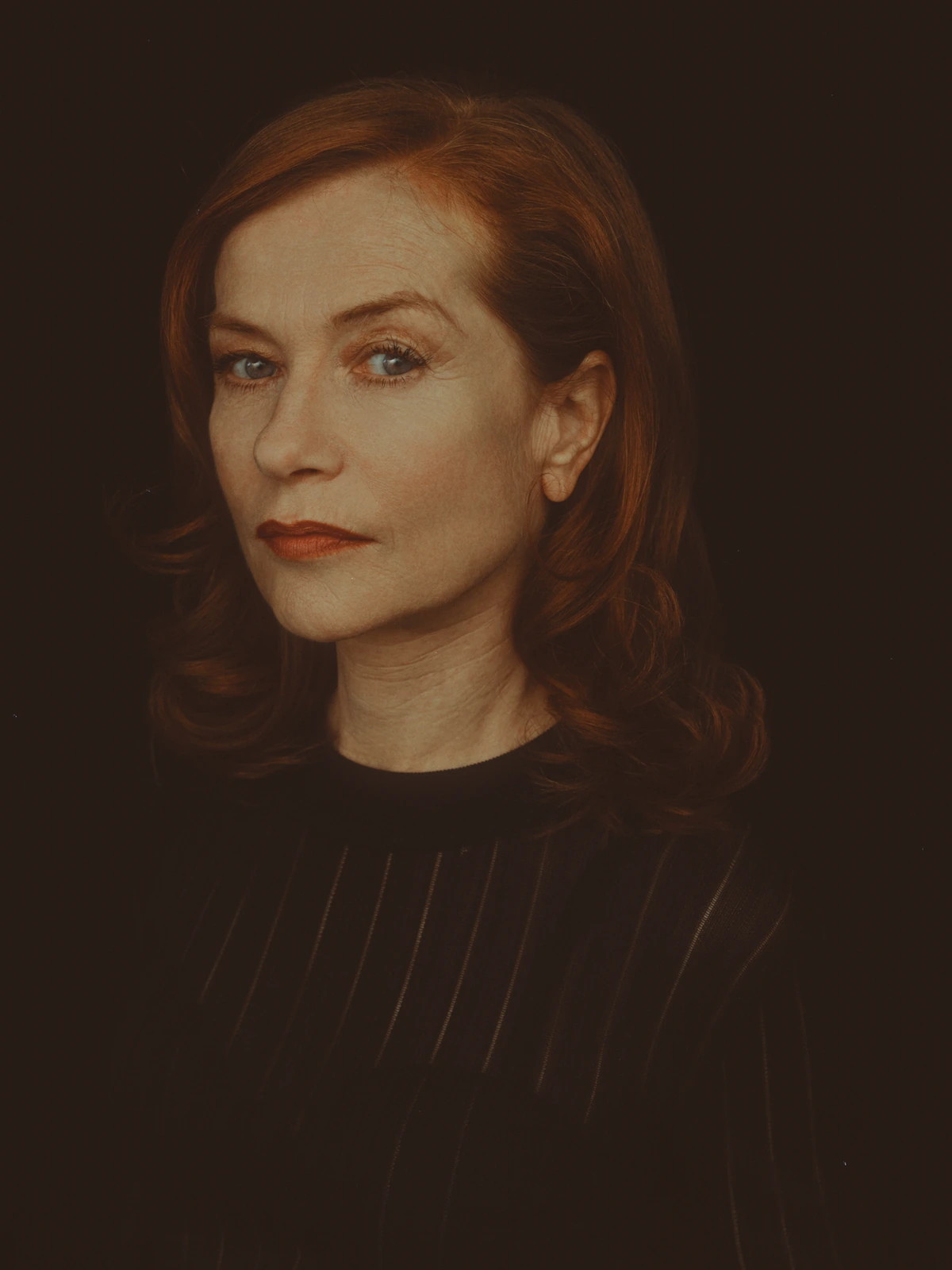 Actress Isabelle Huppert, star of Elle, in a dark striped top against deep shadow, auburn hair and cool gaze in warm amber.
