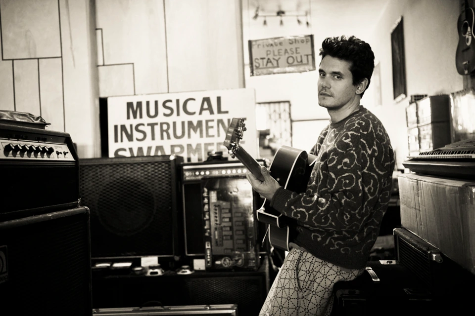 Musician John Mayer holding an acoustic guitar in a vintage instrument shop, patterned sweater, sepia-toned, glancing back.