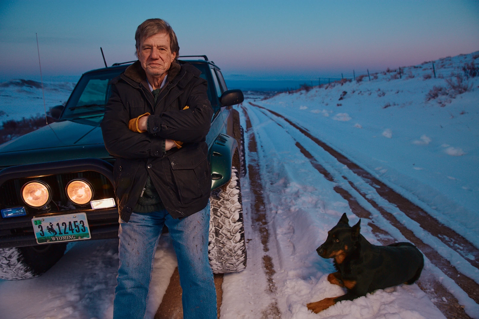Director John McTiernan, arms crossed in a dark jacket, leaning on a Wyoming truck at dusk, black dog at his side on a snowy.